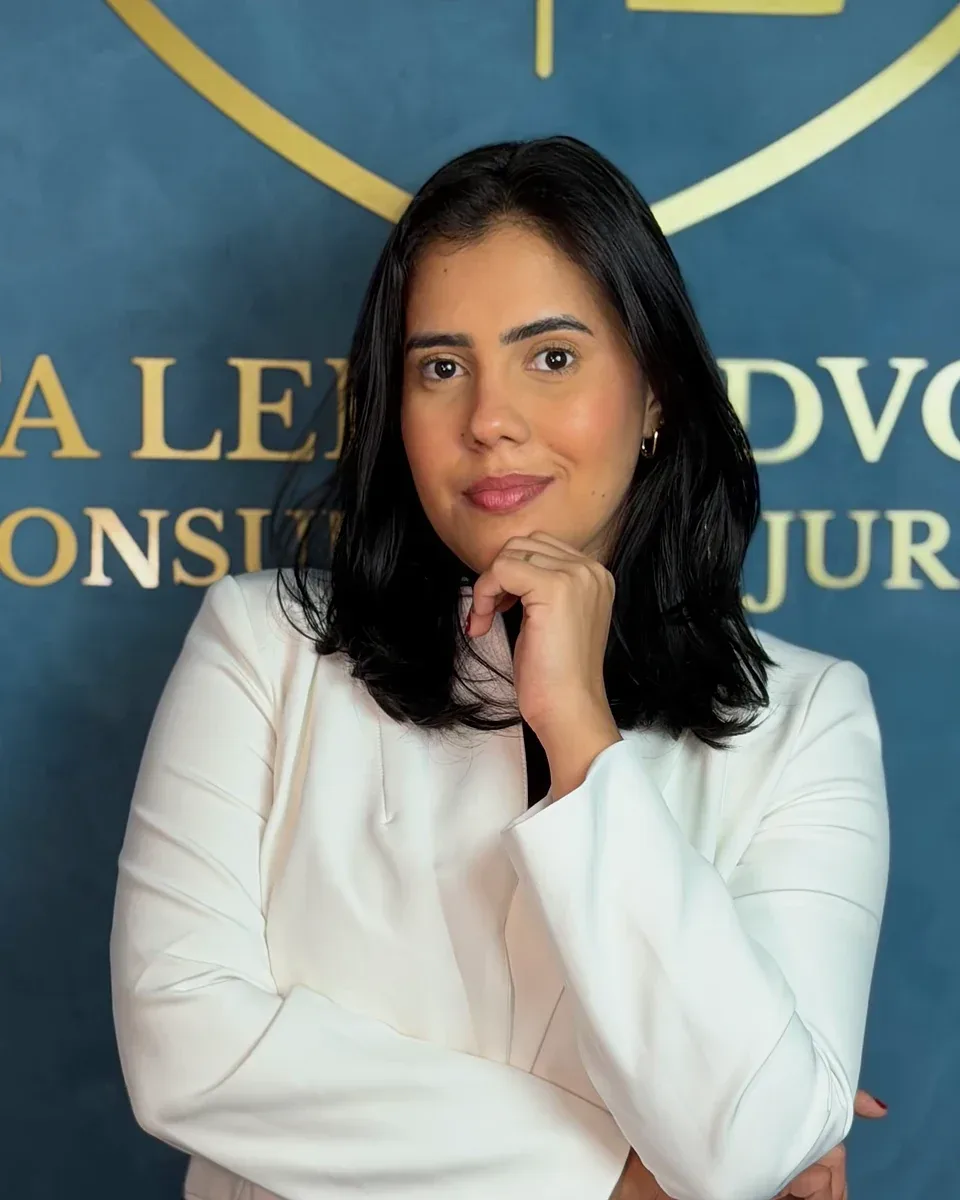 Bel.ª Karen Costa Leite, de blazer branco e com a mão junto ao queixo, diante da parede azul com o brasão dourado do escritório.